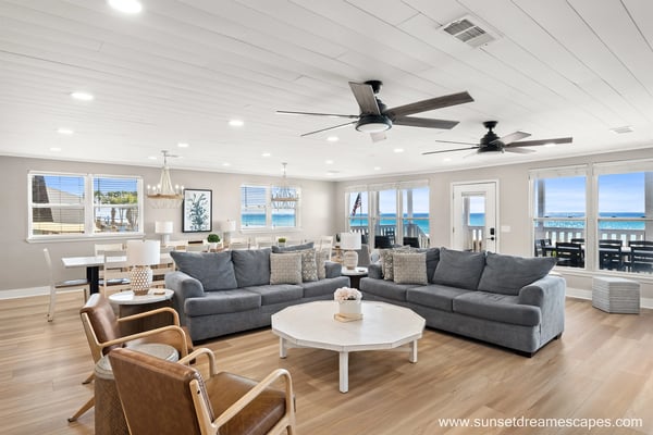 Spacious open-plan living room with gray sectional sofa, ocean view through multiple glass doors, and ceiling fans overhead.