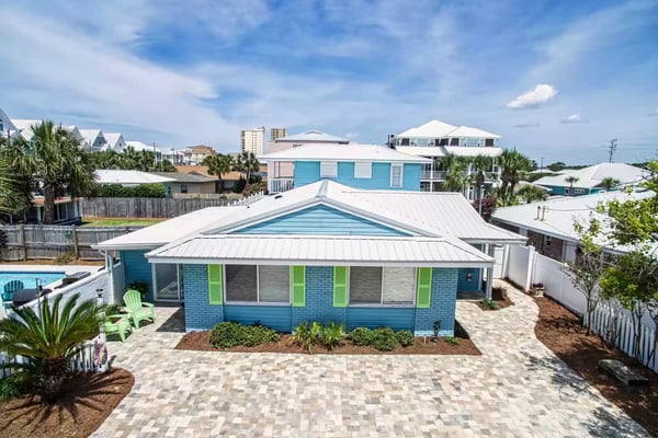 Blue and white beach house with metal roof, paver driveway, and palm trees in coastal neighborhood